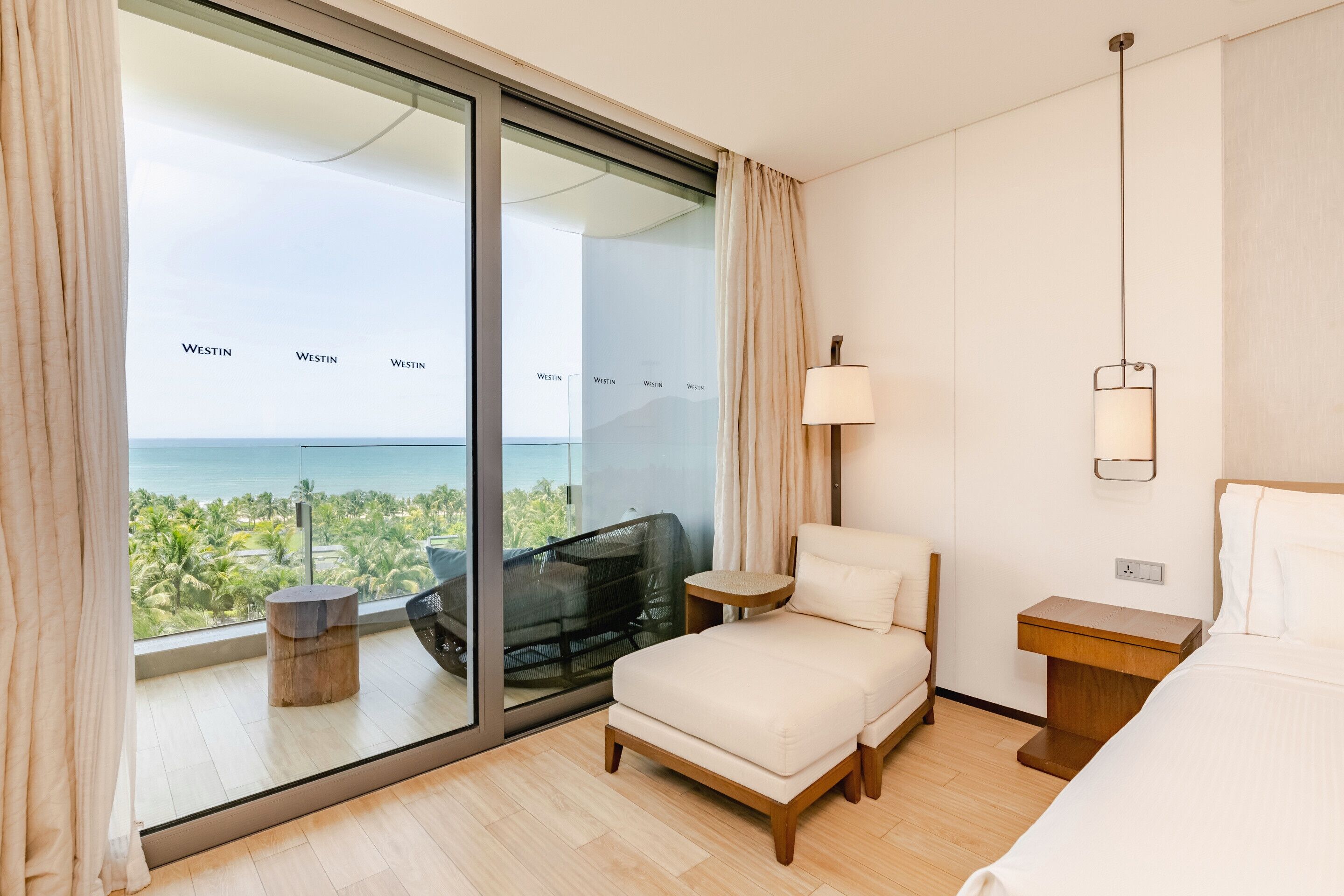 A hotel room with a large sliding glass door leading to a balcony with a view of the ocean. There is a bed, a bedside table, and a chair on the balcony.