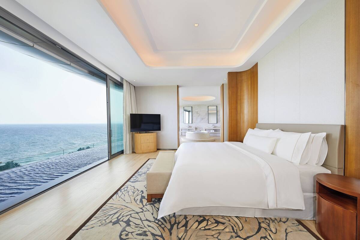 Une chambre d’hôtel moderne dotée d’un grand lit, d’une télévision et d’une salle de bain avec miroir, et offrant une vue sur l’océan.