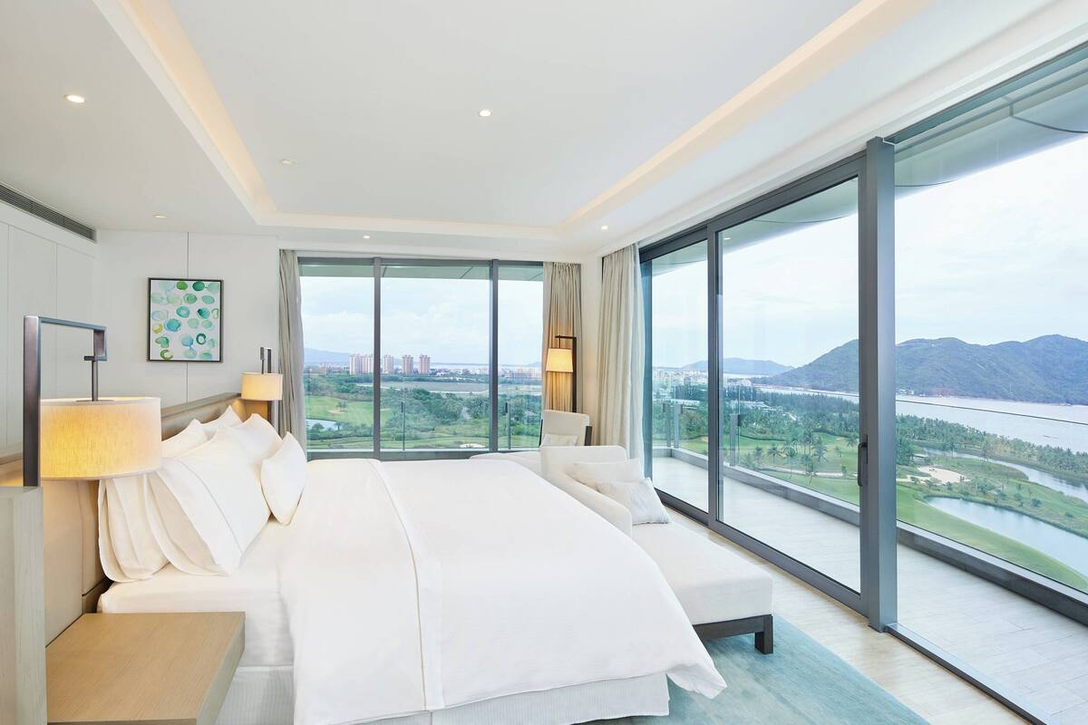 Une chambre d’hôtel moderne avec un grand lit, deux tables de chevet et une vue sur le paysage urbain et les montagnes.