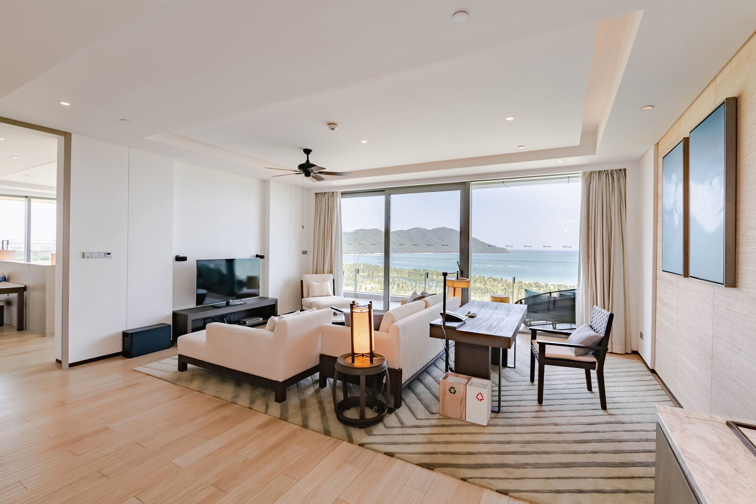 A modern living room with a large TV, white sofas, a coffee table, and a dining area with a view of the sea.