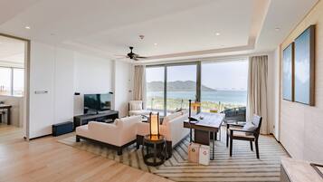 A modern living room with a large TV, white sofas, a coffee table, and a dining area with a view of the sea.