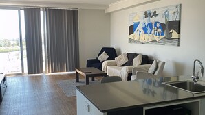 A modern kitchen with a built-in microwave, a dining area with a table and chairs, and a living area with a sofa and a painting on the wall. - Curtis Central Apartments (Gladstone)