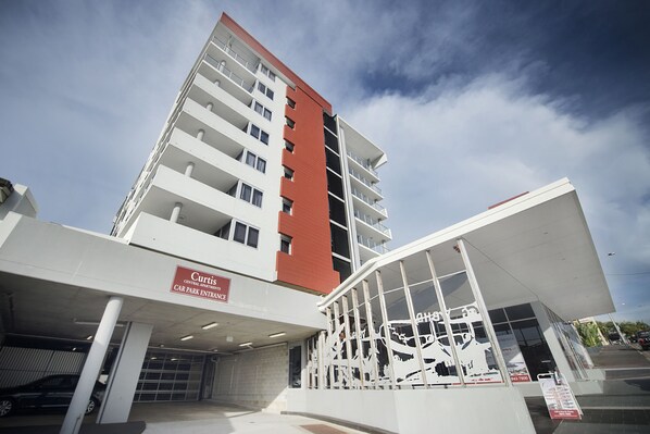 Un edificio de varios pisos con fachada roja y blanca, un letrero que indica la entrada a un estacionamiento y una entrada moderna de vidrio.