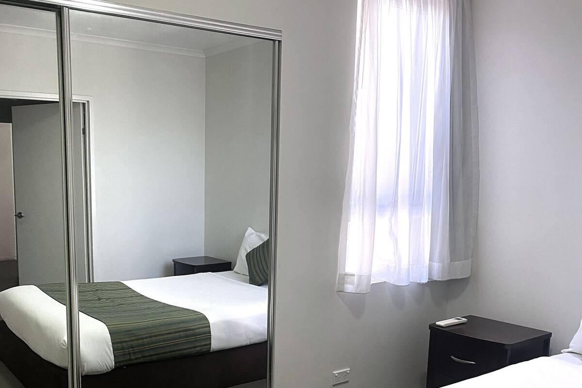 A hotel room with a bed, a bedside table, a nightstand, a mirror, and a window with curtains.