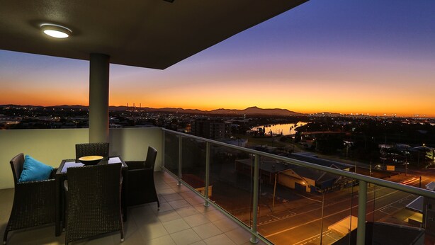 A balcony with wicker furniture and a view of a cityscape at dusk. - Curtis Central Apartments (Gladstone)