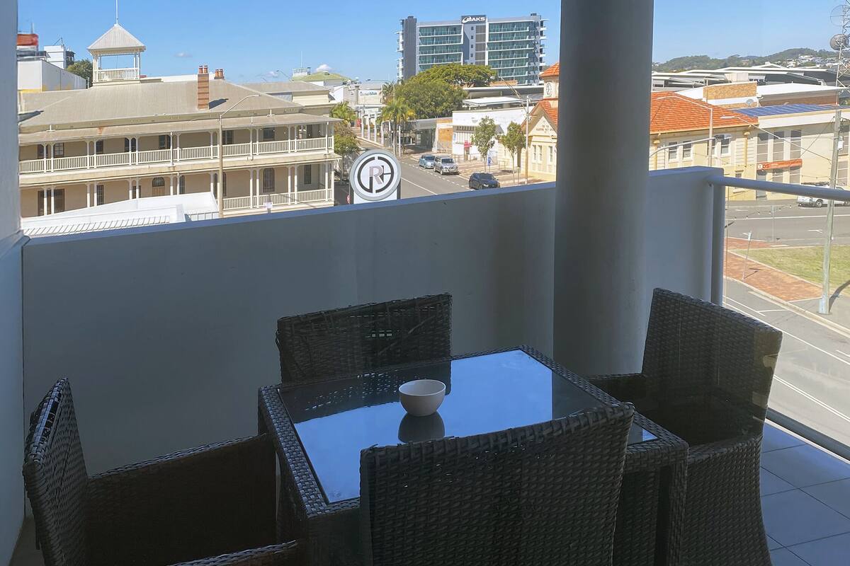 A balcony with wicker furniture and a table, overlooking a cityscape with buildings and a clear sky.