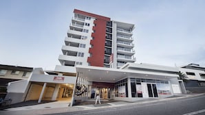 A multi-story hotel with a modern design, featuring a prominent red and white facade, a covered entrance, and a visible car park. - Curtis Central Apartments (Gladstone)