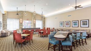A spacious dining area with high ceilings, multiple seating arrangements including booths and tables, and a mix of red and blue upholstered chairs. - Homewood Suites By Hilton San Bernardino (San Bernardino)