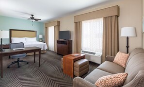 Habitación de hotel con cama, escritorio, silla, sofá, televisor y ventanas con cortinas.