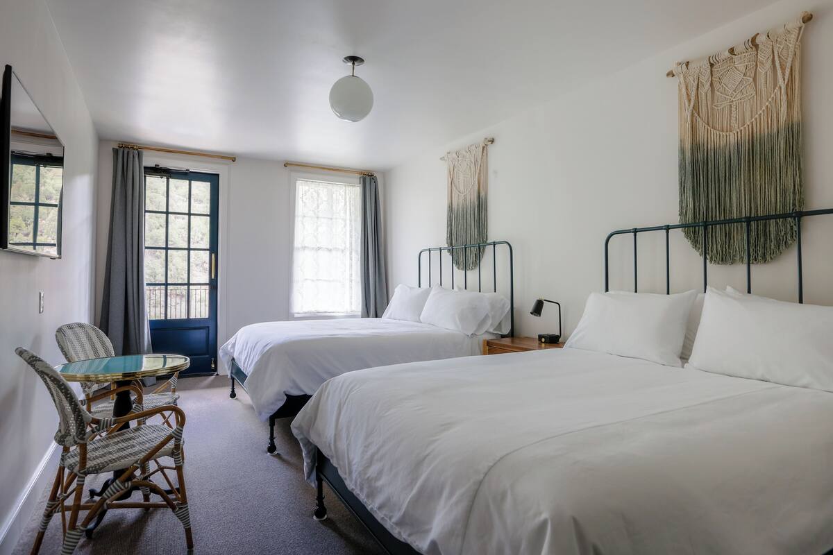 Een kamer met twee bedden, een tafel met stoelen, een spiegel en een wanddecoratie van macramé.