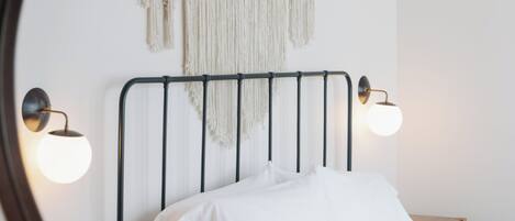 Una cama con ropa de cama blanca, un tapiz de macramé y dos luces fijadas en la pared.