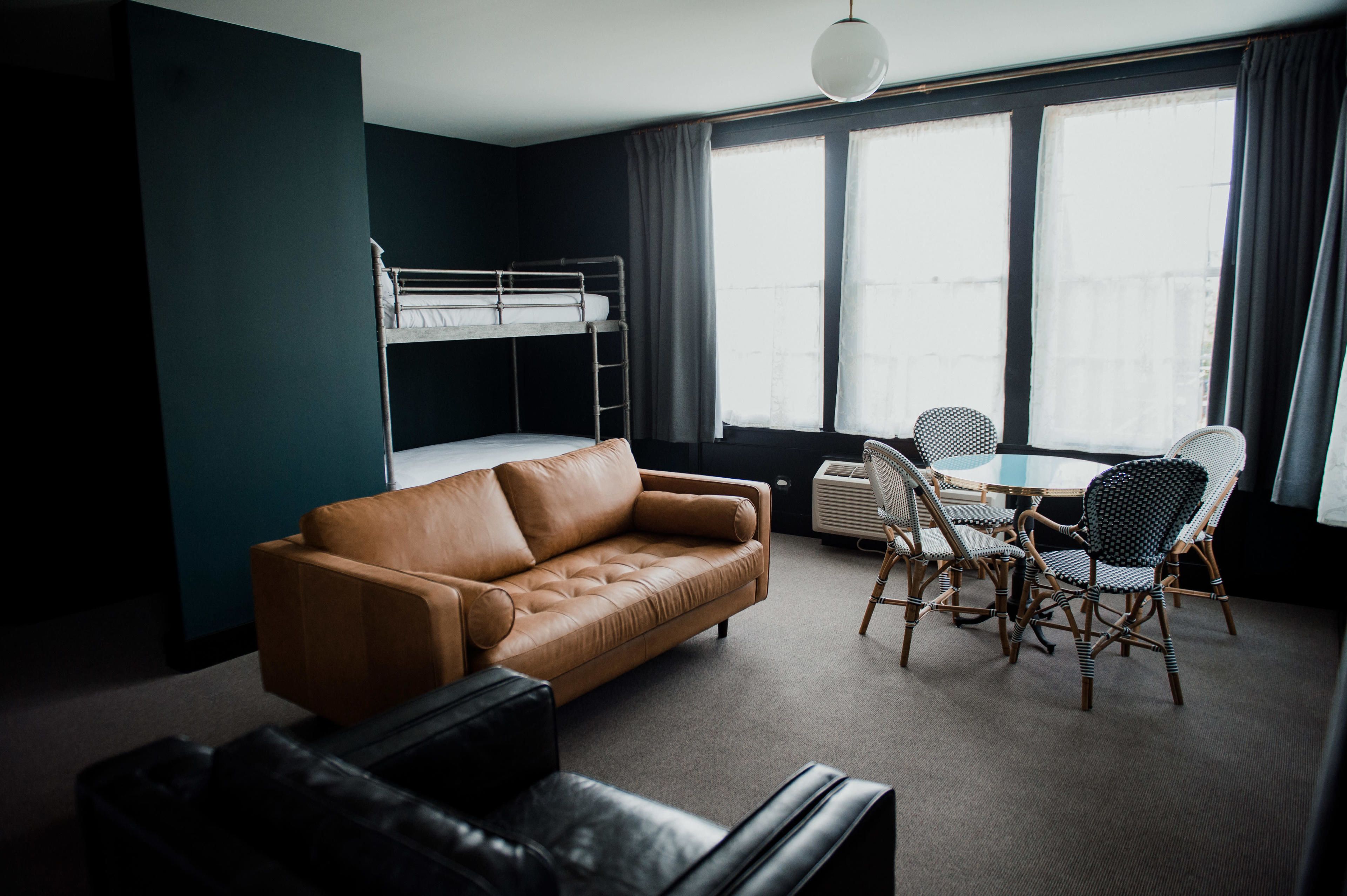A room with a bunk bed, a leather sofa, a dining table with chairs, and large windows with curtains.