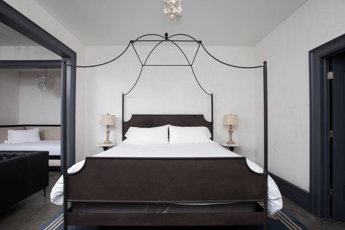 Een hemelbed met witte lakens, twee bedlampjes en een kroonluchter.