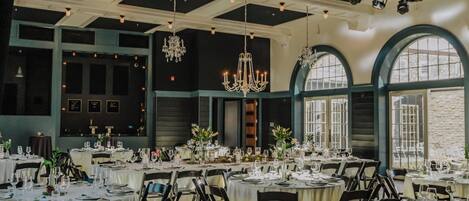 A spacious dining area with multiple round tables set for a formal event, featuring chandeliers and large windows.