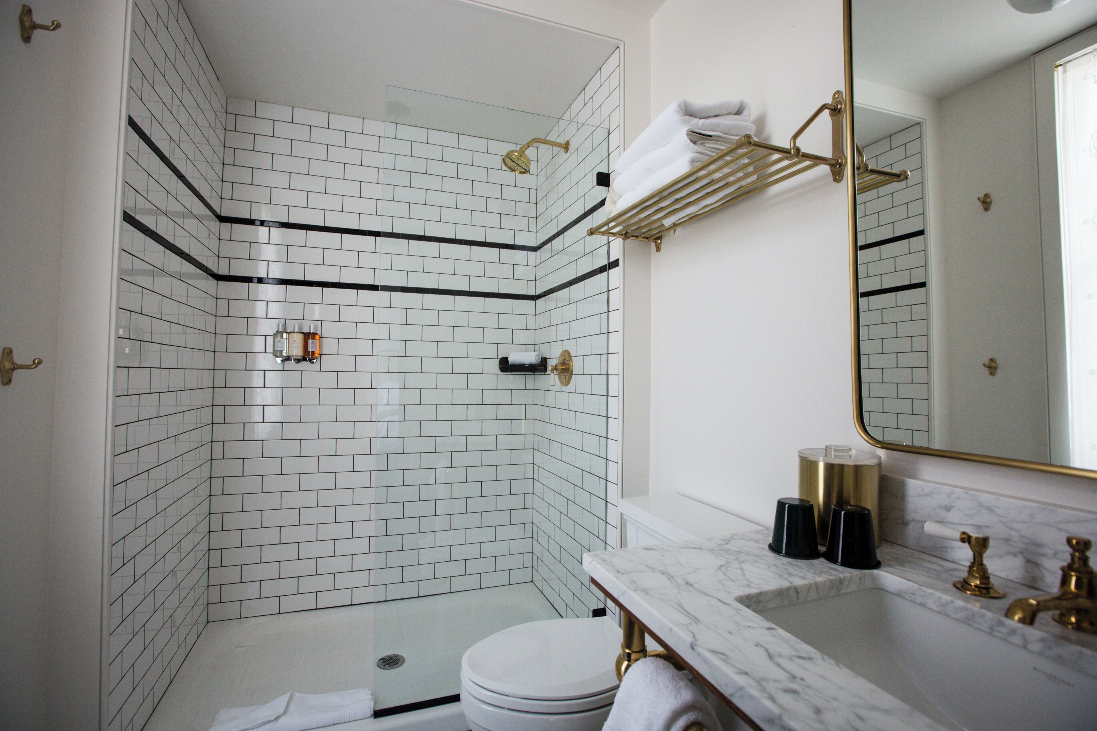Un baño con azulejos blancos tipo metro, una cabina de ducha de vidrio, inodoro blanco, lavamanos de mármol y un espejo con marco dorado.