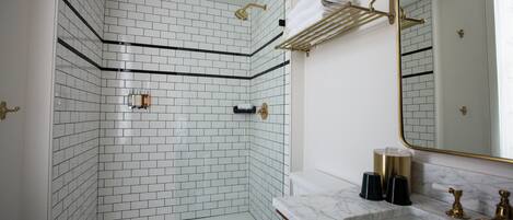 Un baño con azulejos blancos tipo metro, una cabina de ducha de vidrio, inodoro blanco, lavamanos de mármol y un espejo con marco dorado.