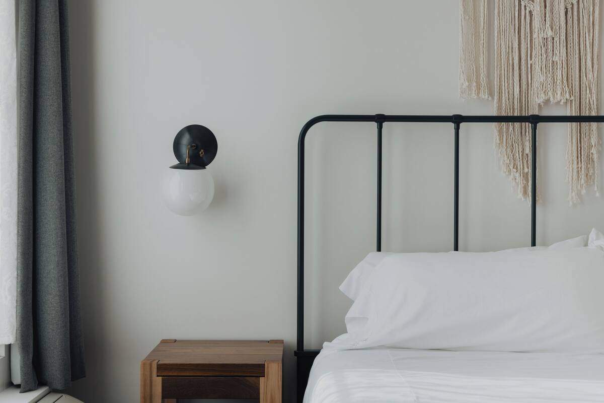 Een slaapkamer met een bed, nachtkastje, een wandlamp en een macramé wanddecoratie.