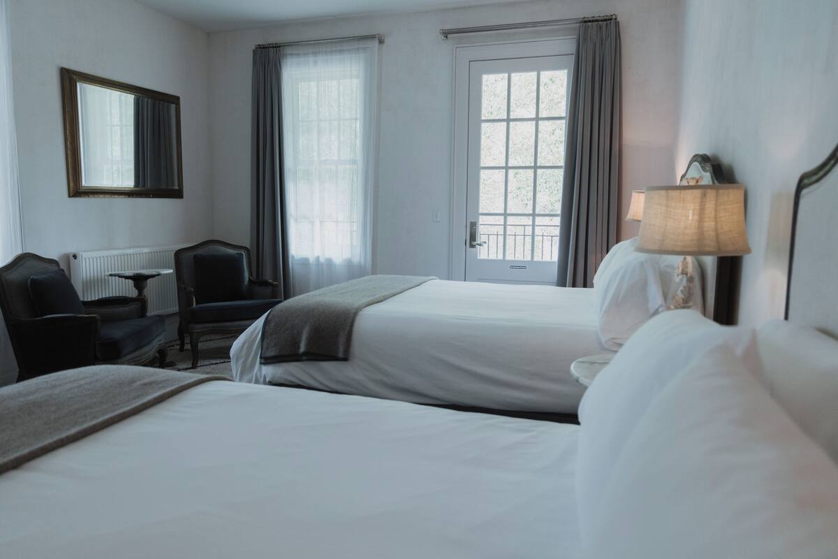 Een hotelkamer met twee bedden, een bureau, een stoel, een lamp en een raam met gordijnen.