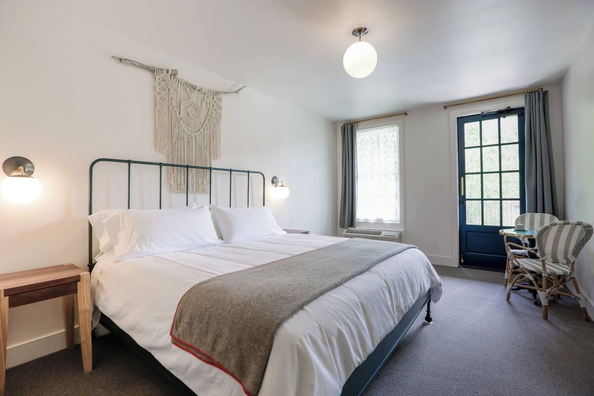 Een slaapkamer met een bed, nachtkastjes, een stoel, een raam met gordijnen en een wanddecoratie van macramé.