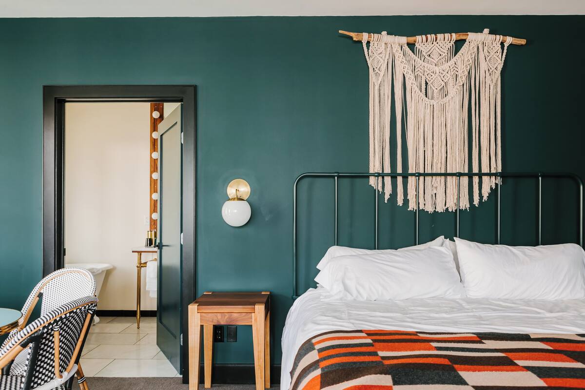 Een slaapkamer met een bed, een stoel, een klein tafeltje, wanddecoratie van macramé en een kaptafel met spiegel en verlichting.