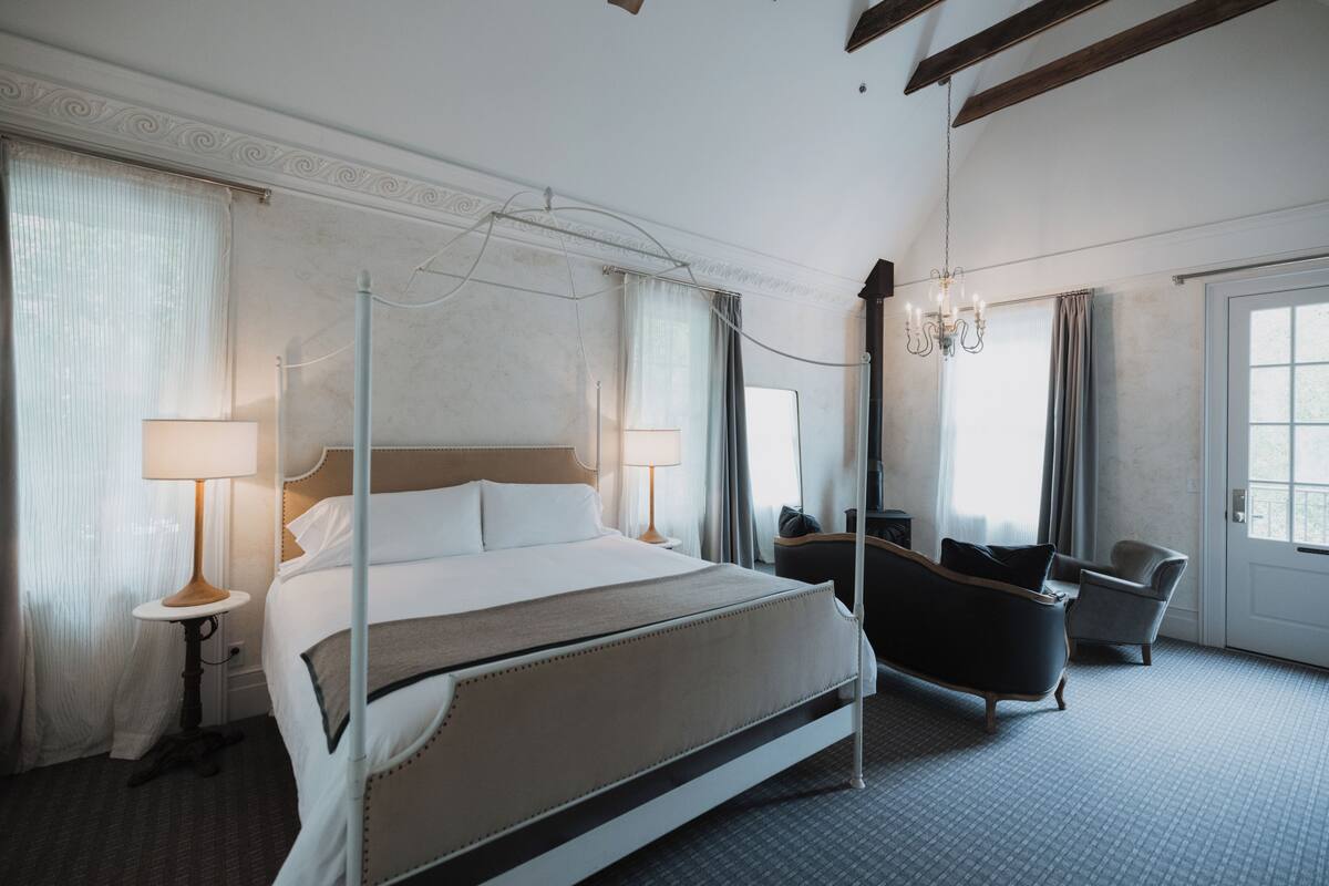 Een hotelkamer met een hemelbed, twee bedlampen, een kroonluchter, een zithoek met een fauteuil en een bank, en een groot raam.