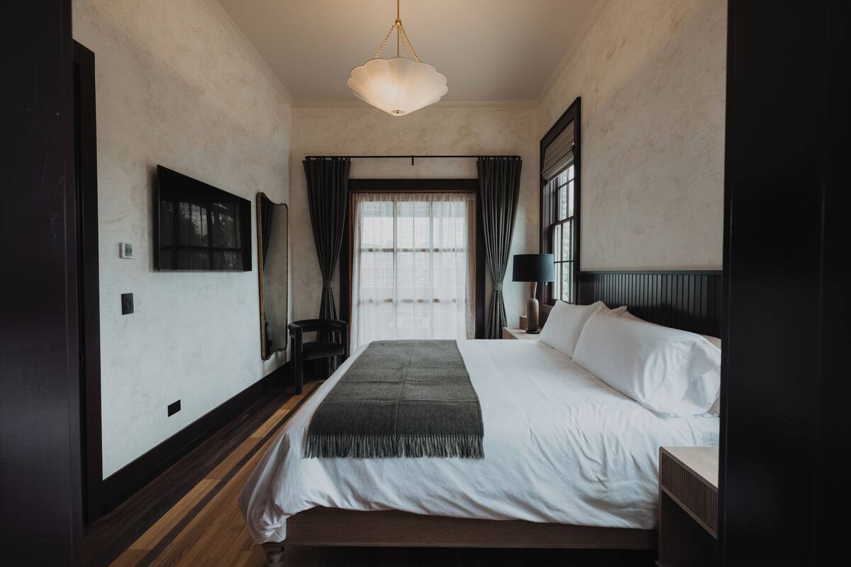 Een slaapkamer met een bed, een hanglamp, een raam met gordijnen en een aan de muur bevestigde televisie.
