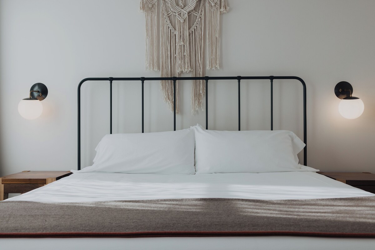 Una cama con sábanas blancas, un colgador de macramé en la pared y dos lámparas de pared.