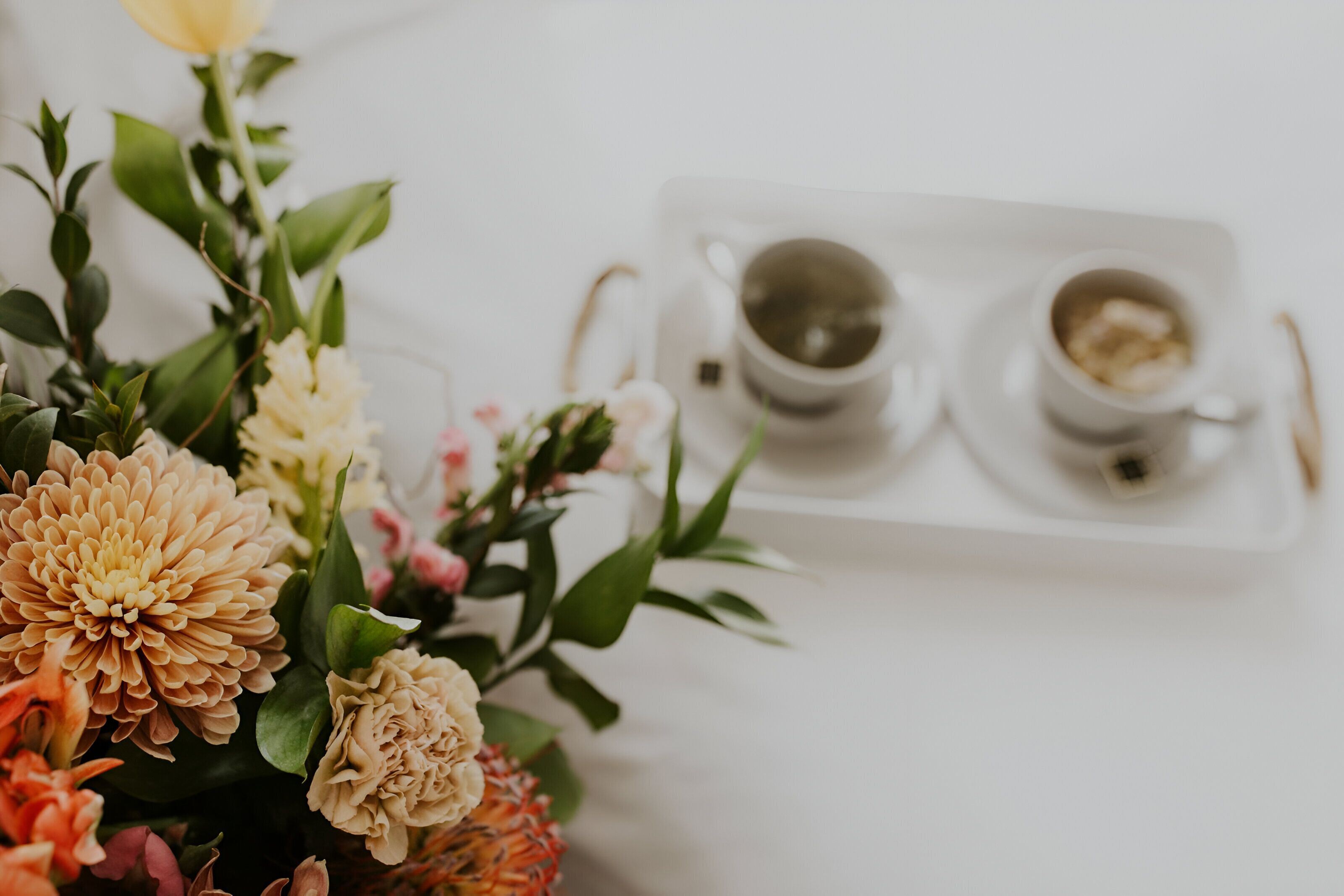 A white table with a floral arrangement and two white cups with saucers, one containing a beverage and the other with a dessert.