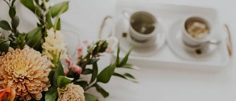 Una mesa blanca con un arreglo floral y dos tazas blancas con platillos, una con una bebida y la otra con un postre.