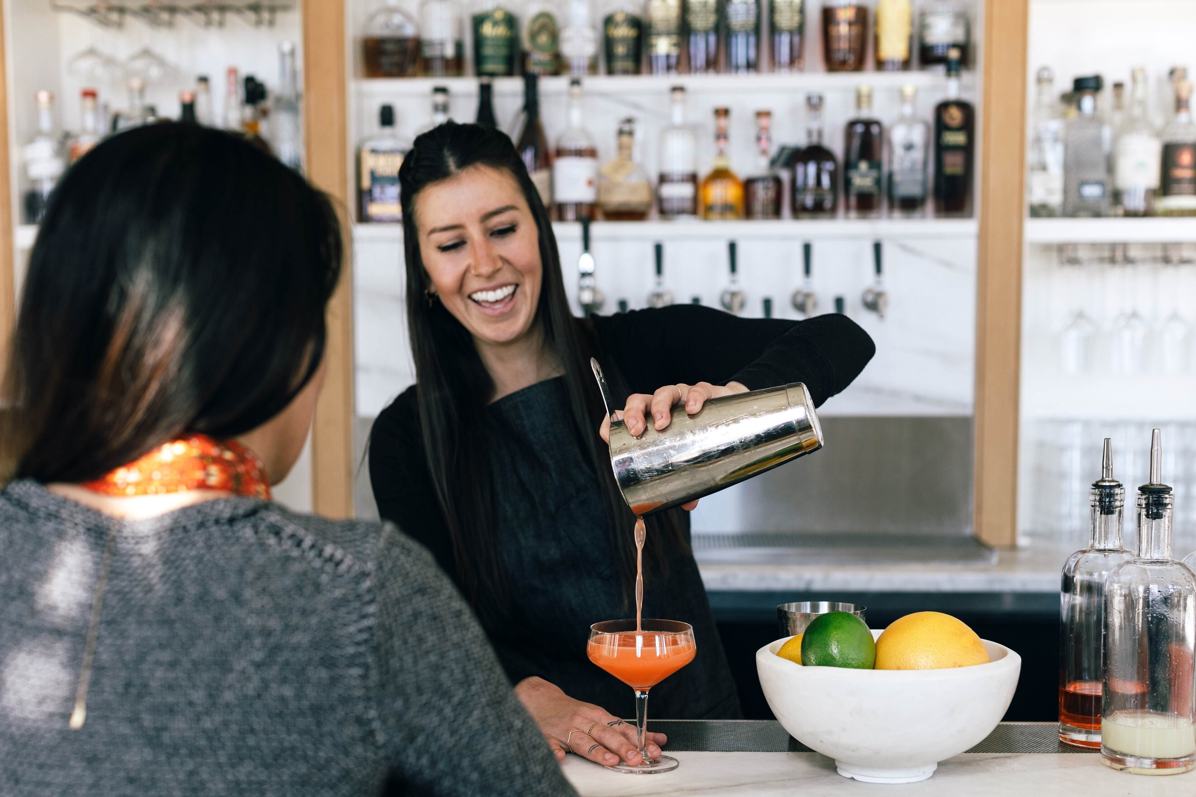 Un barman verse une boisson dans un verre à martini dans un bar où l’on peut voir une variété de bouteilles en arrière-plan.