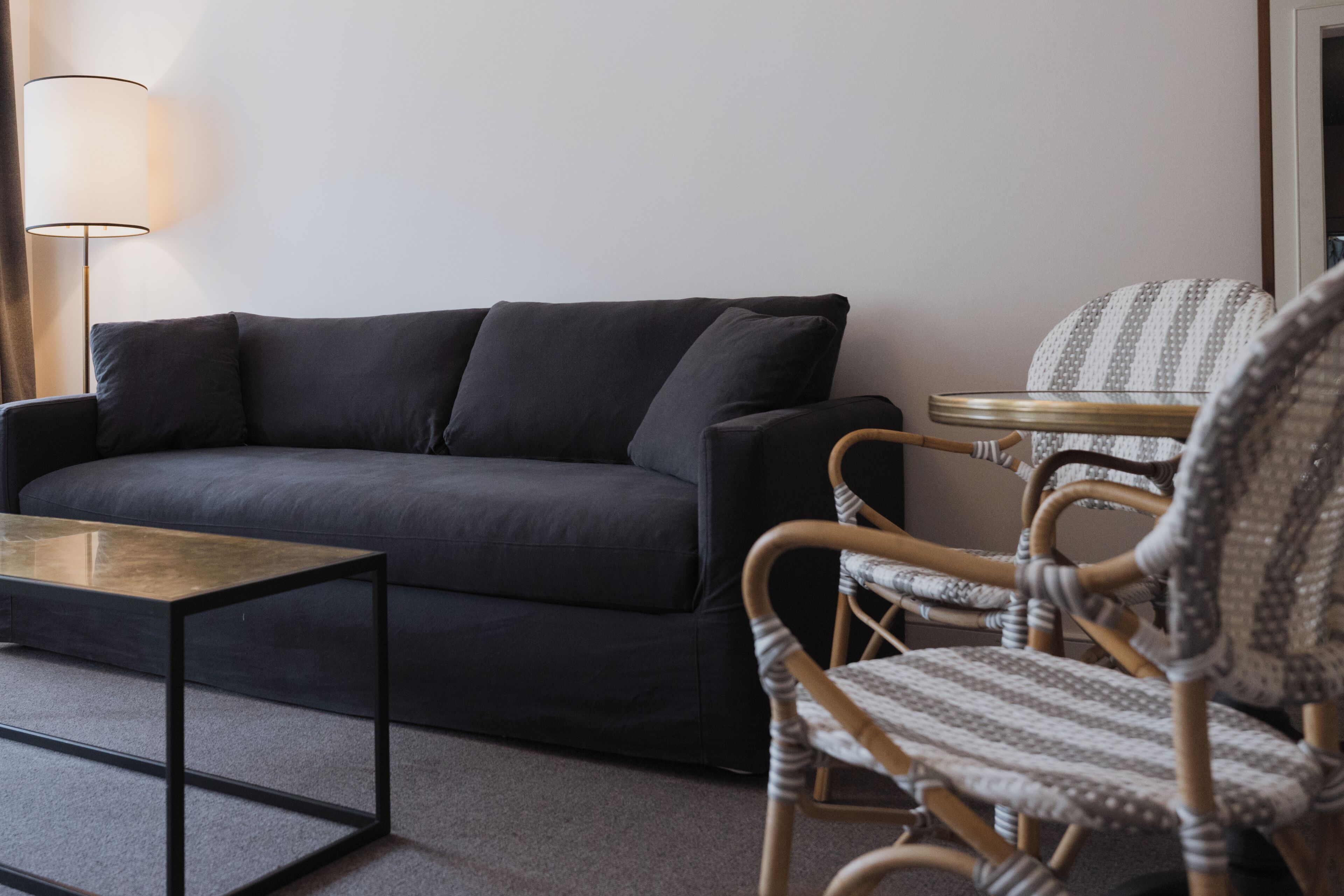 A dark-colored sofa, a small round table, and a wicker chair in a room with a plain wall and a floor lamp.