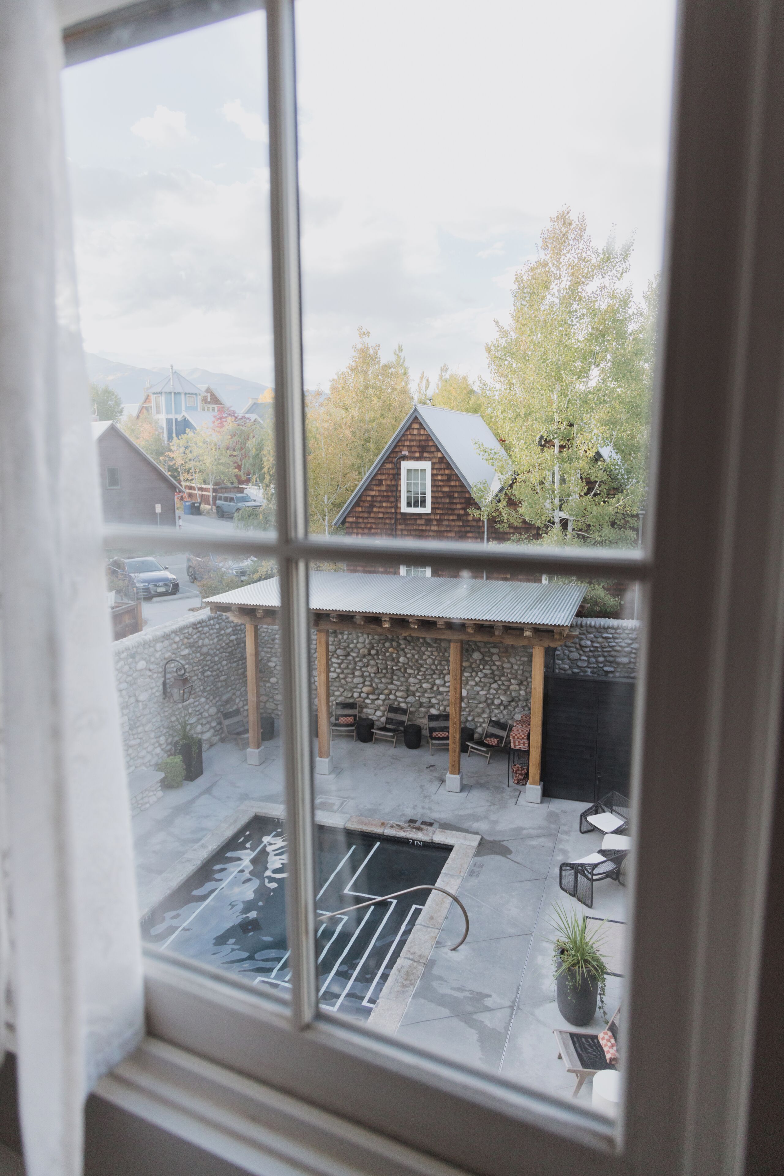 A view through a window of a stone patio area with outdoor seating, a small pool, and a wooden building in the background.