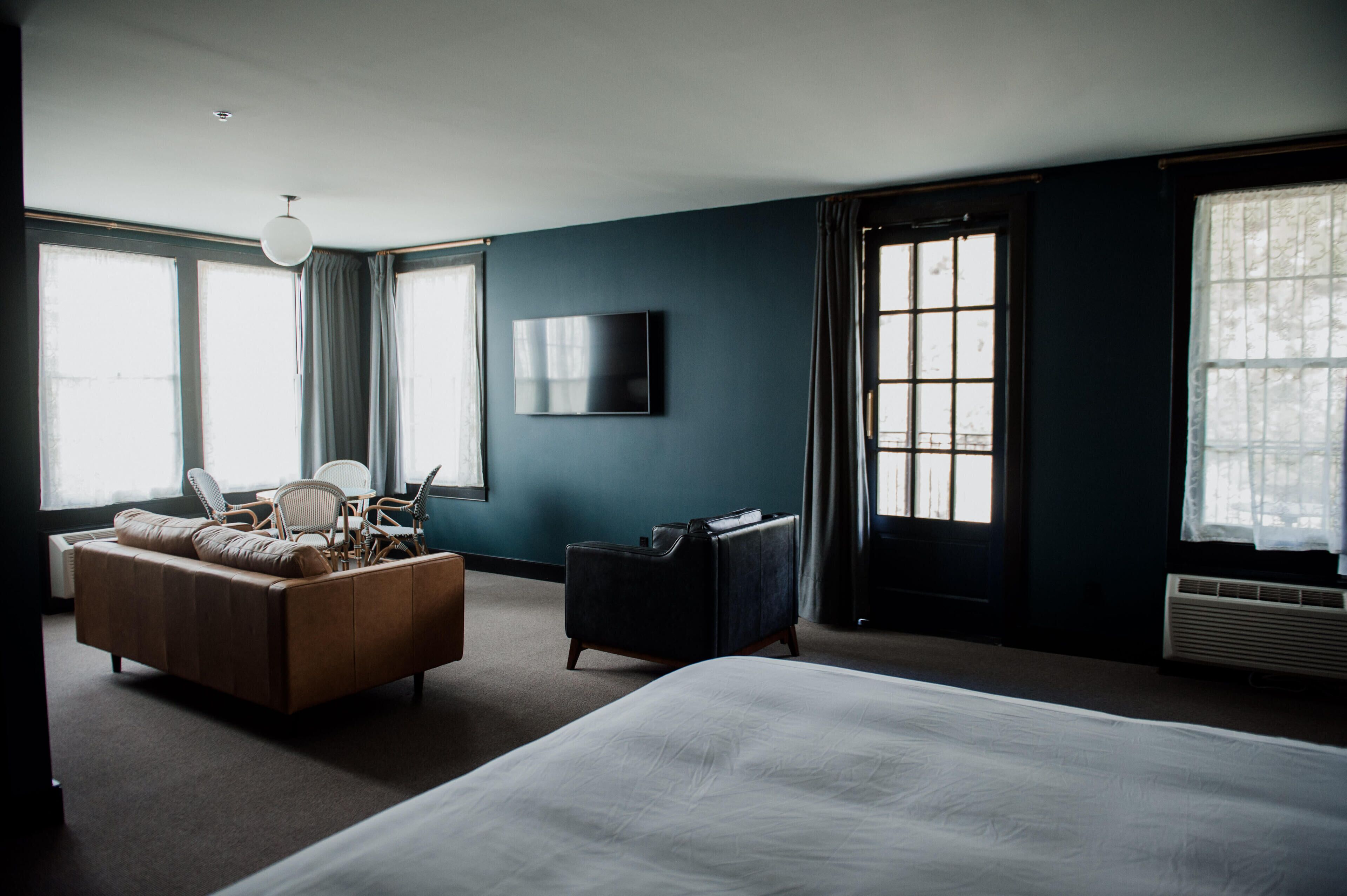 A hotel room with a large bed, a seating area with a sofa and chairs, a flat-screen TV mounted on the wall, and large windows with curtains.