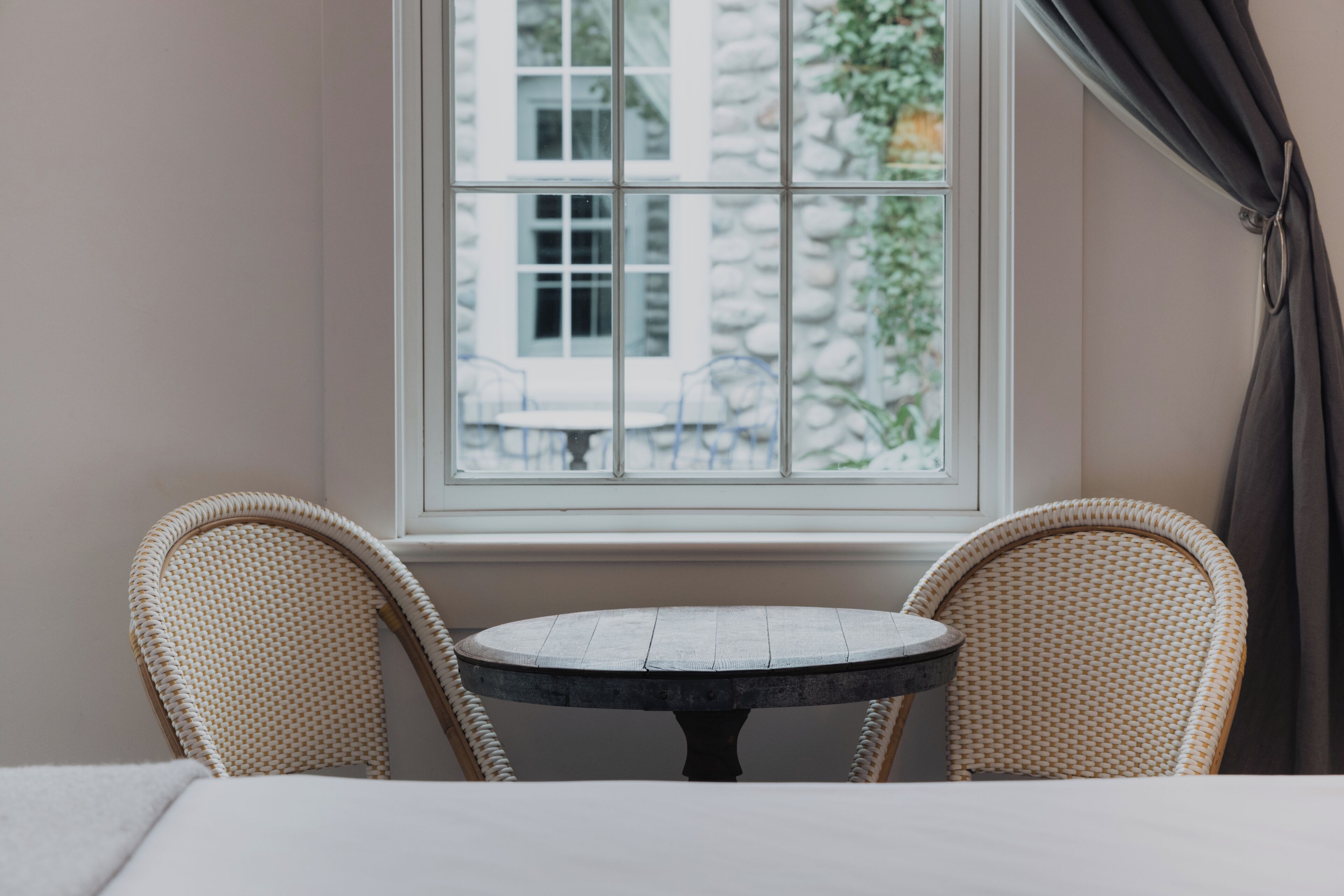 Ein Zimmer mit einem runden Tisch und zwei Korbstühlen, ein Fenster mit Blick auf ein Gebäude sowie ein Vorhang auf der rechten Seite.