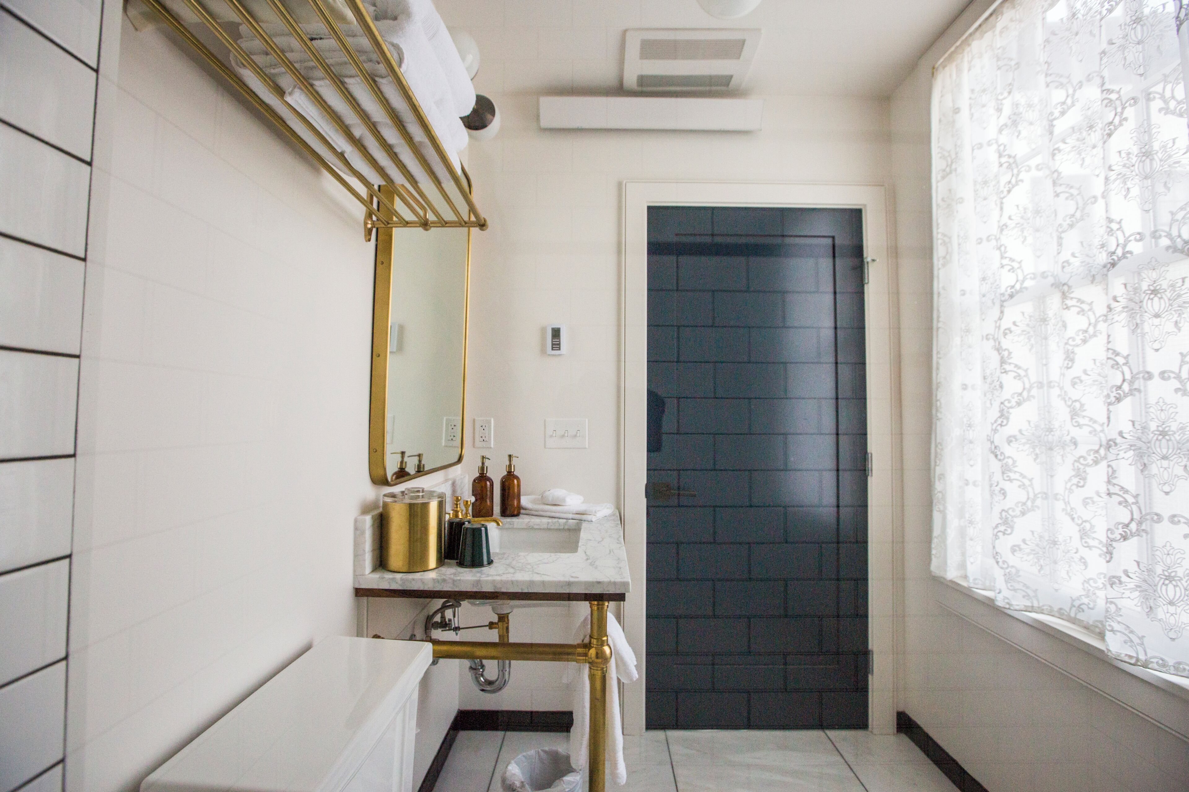 A bathroom with a white vanity, a gold-rimmed mirror, a dark door, and a window with patterned curtains.