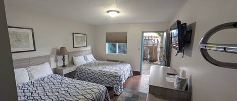 A hotel room with two beds, a desk, a chair, a TV, and a window with a view of a staircase.