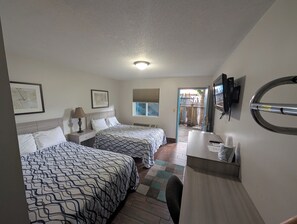 A hotel room with two beds, a desk, a chair, a TV, and a window with a view of a staircase.