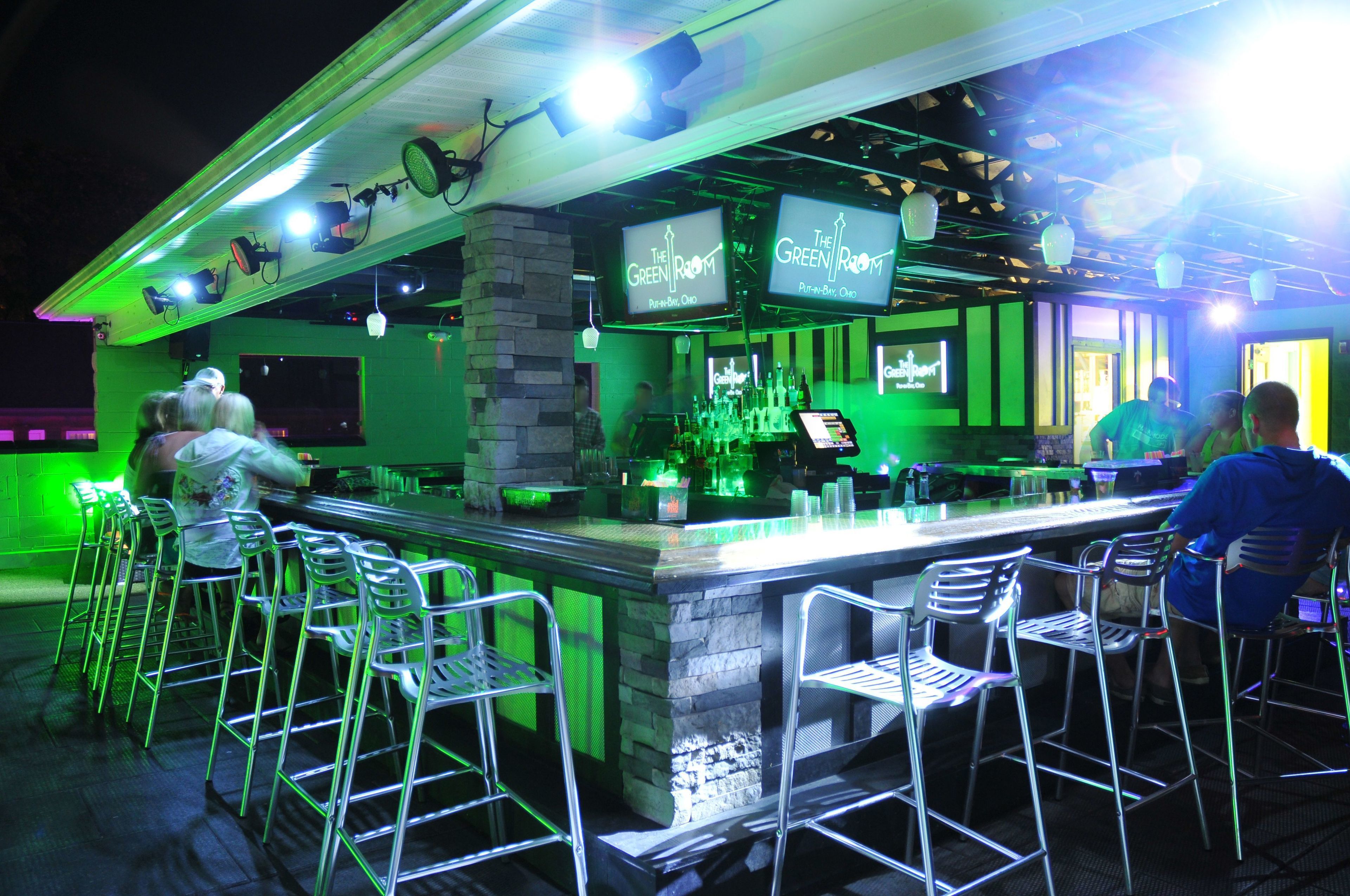 An outdoor bar area with a green-lit bar counter, multiple bar stools, and overhead lighting.