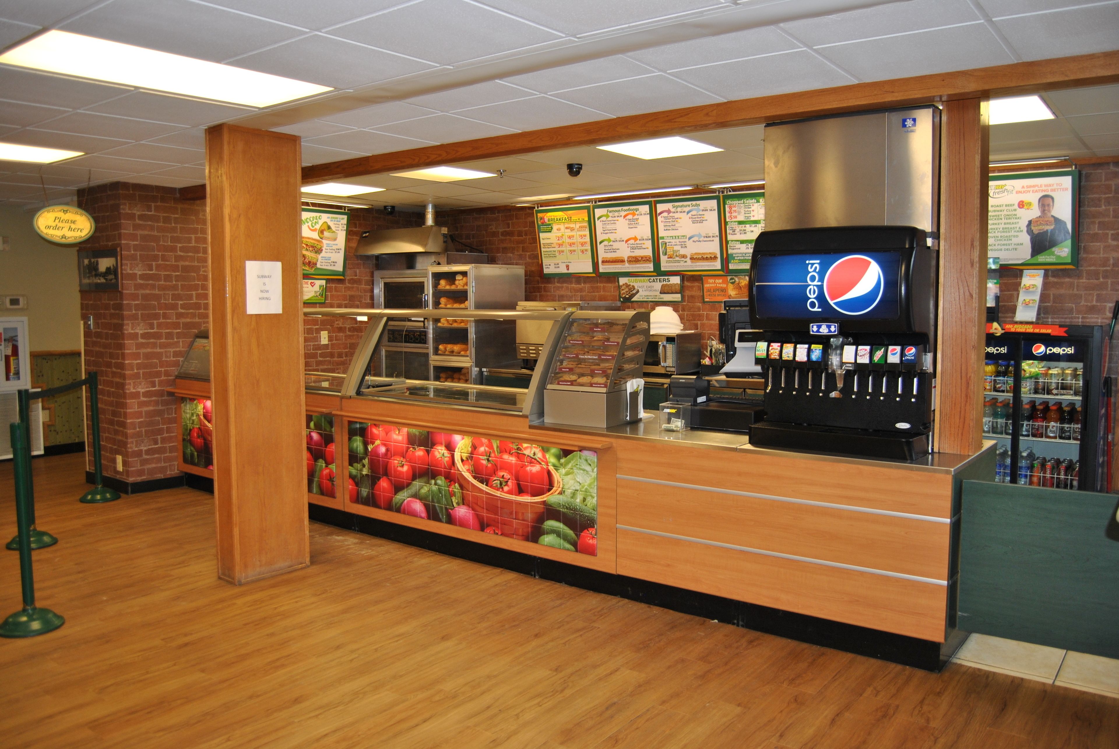 A food service area with a brick wall, a wooden counter, a Pepsi vending machine, and menu boards.