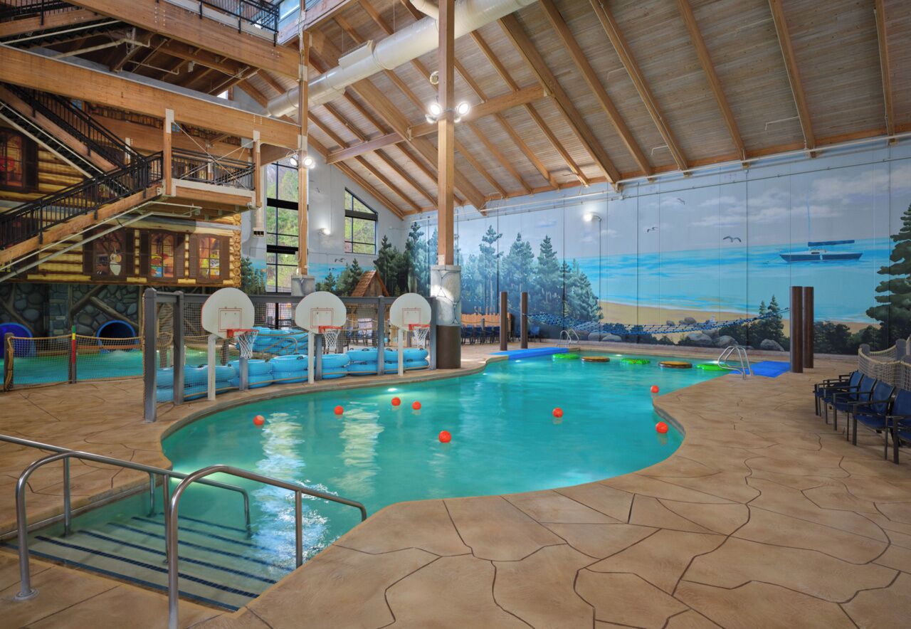 An indoor swimming pool with a mural of a beach scene, wooden beams, and a basketball court.