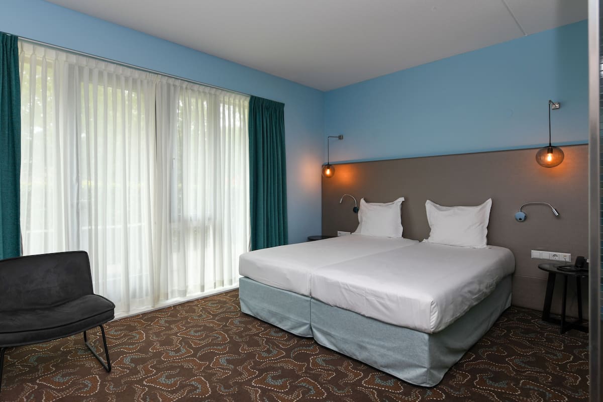 Quarto de hotel com cama, cadeira, mesa de cabeceira e janela com cortinas.