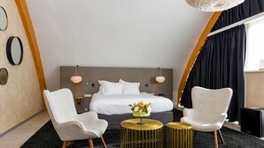 Una habitación de hotel moderna con una cama grande, dos sillas, una mesita con un florero y un ventanal con cortinas.