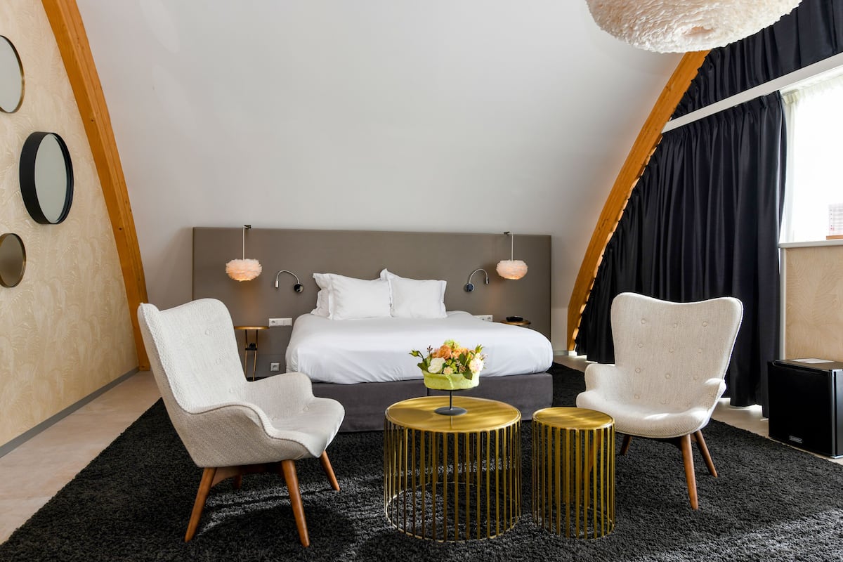 Quarto de hotel moderno com uma cama grande, duas cadeiras, uma mesinha com um vaso e uma janela ampla com cortinas.