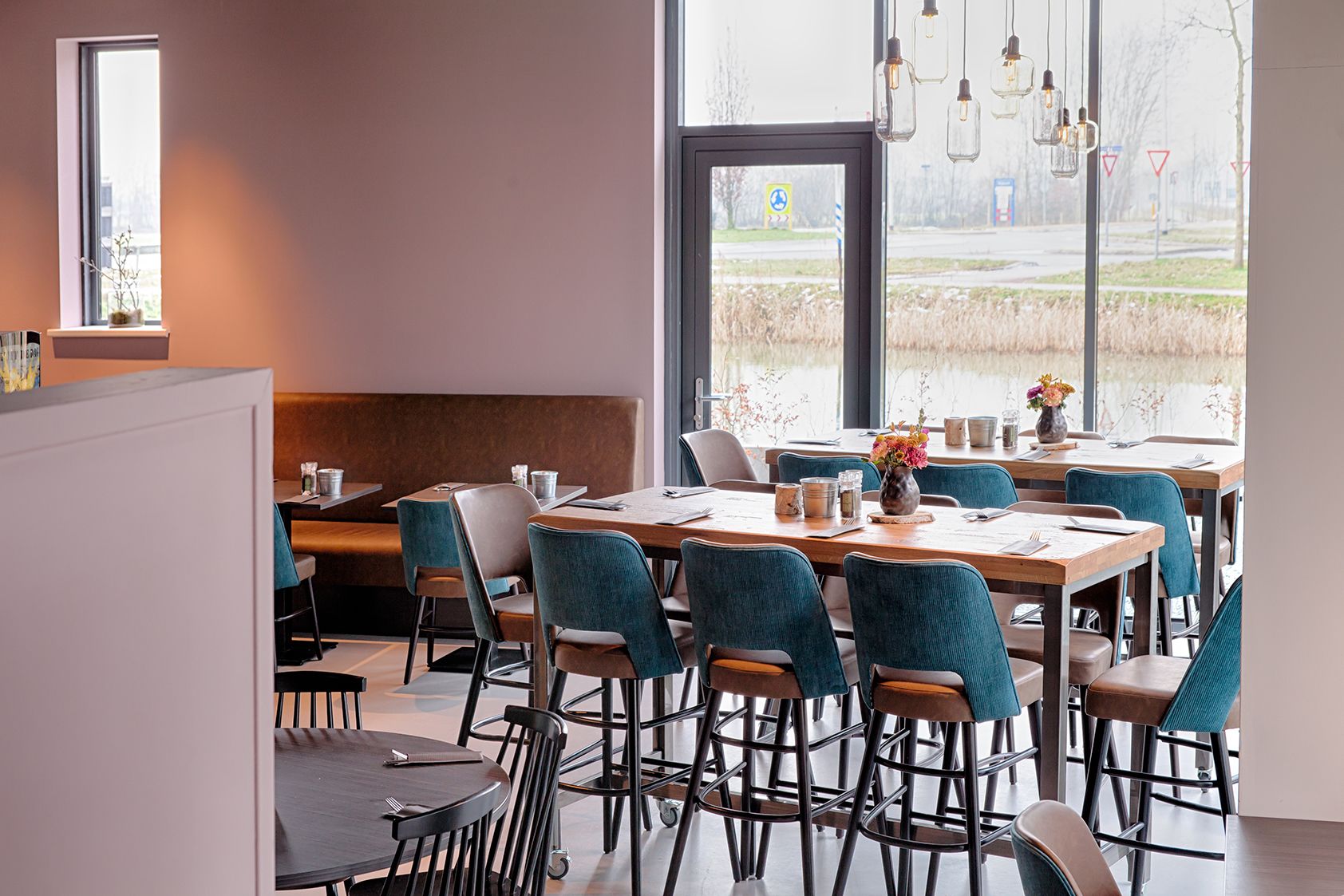 Un espace repas moderne avec des tables et des chaises hautes, un banc et une vue sur une route et des arbres, le tout visible à travers de grandes fenêtres.