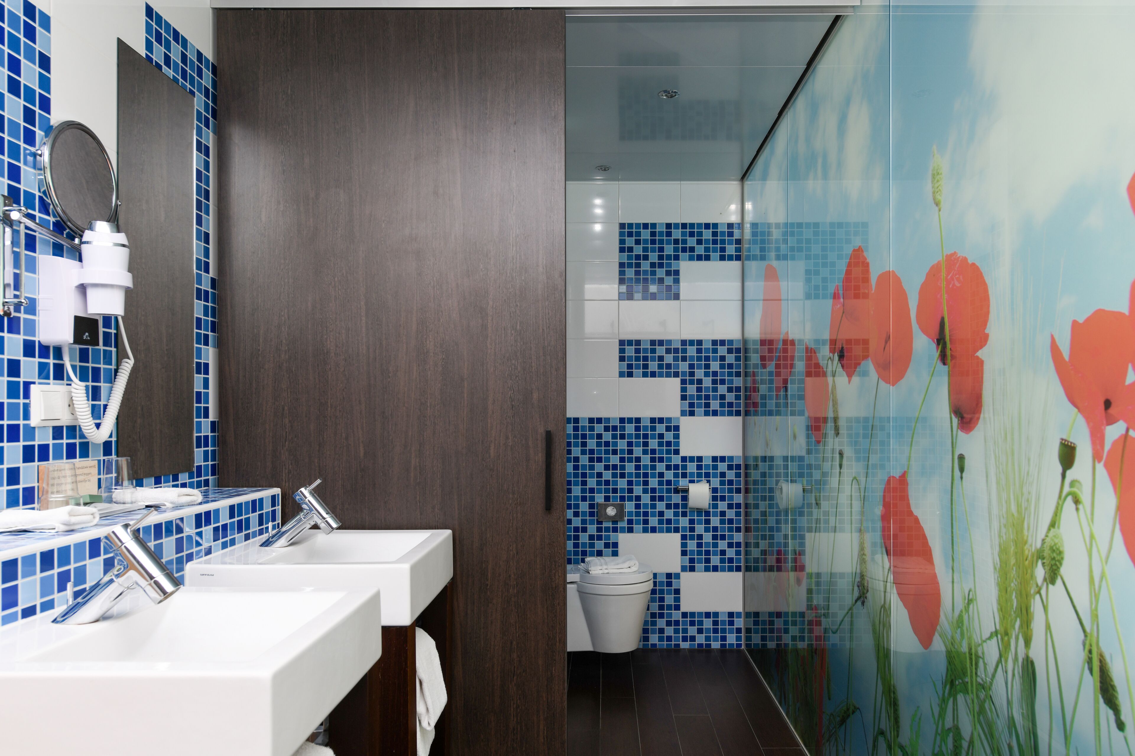 A bathroom with two sinks, a mirror, and a toilet. The walls are tiled with blue and white patterns, and there is a floral mural on the right wall.