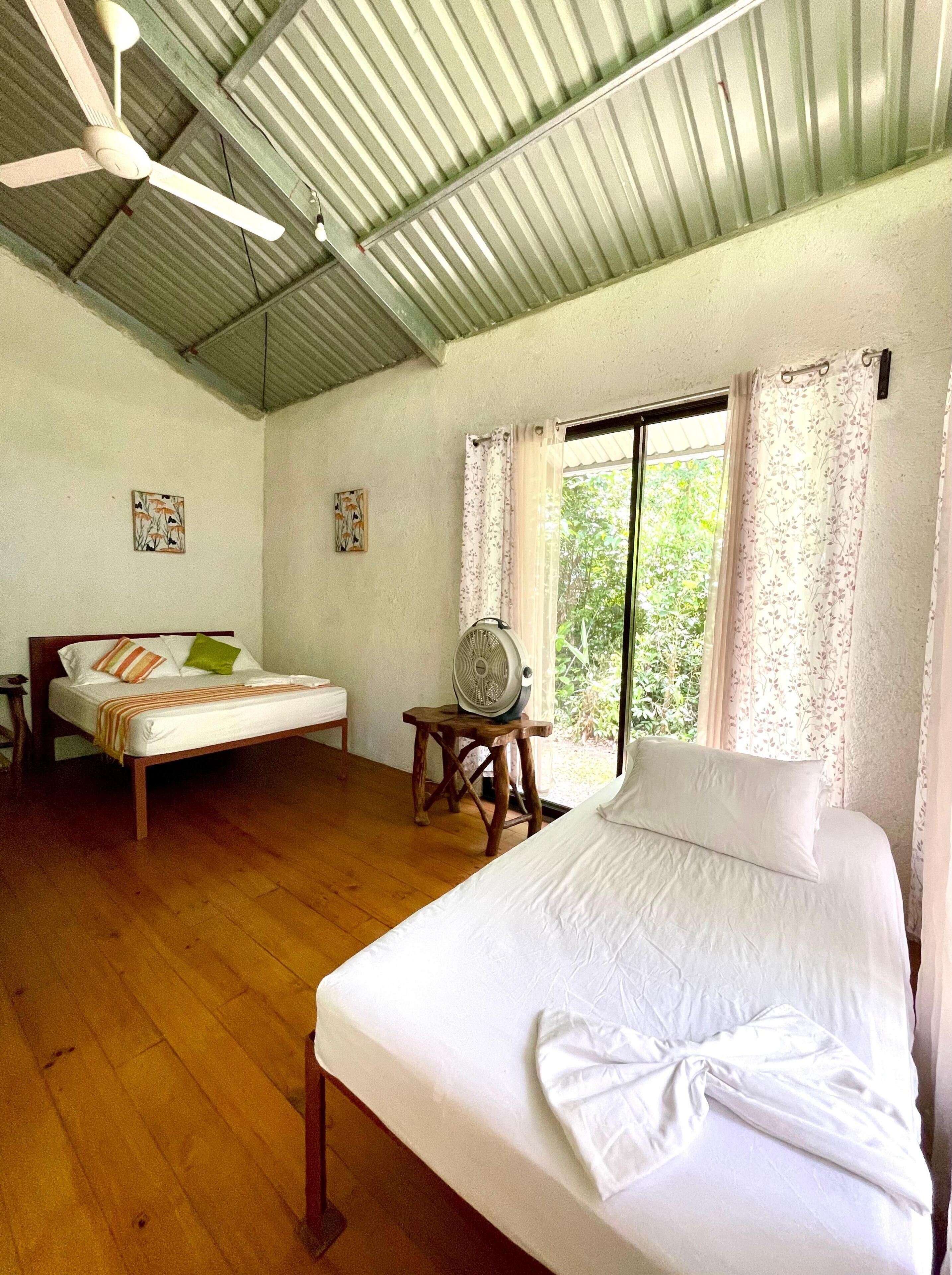 A room with a bed, a smaller bed, a standing fan, and a window with curtains.