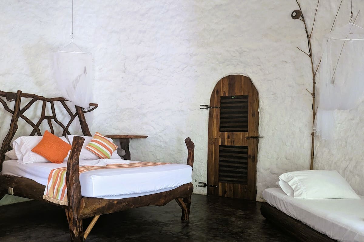 A rustic bedroom with a wooden bed, a small bedside table, a wooden door, and a white wall.