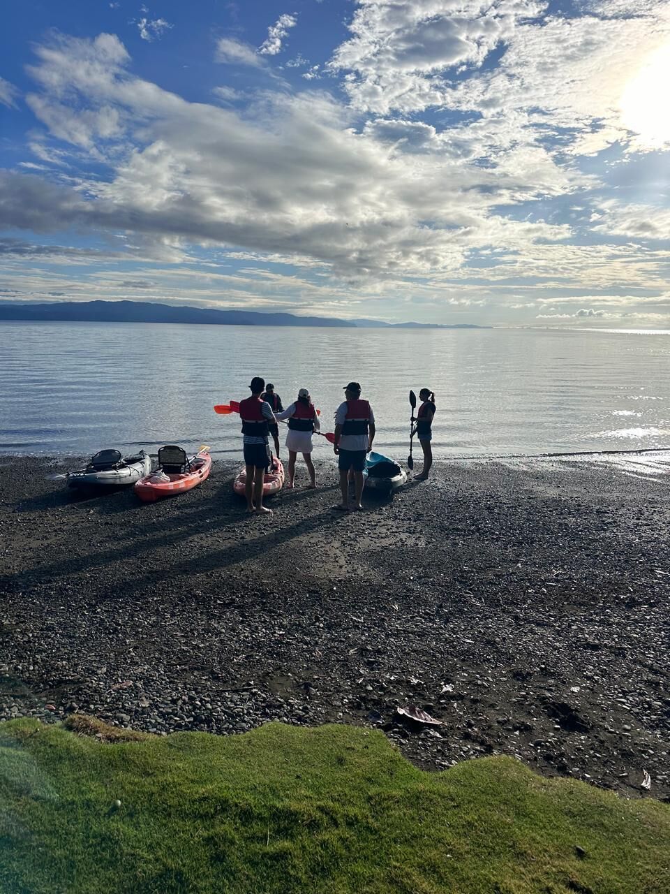 A group of people on a pebbly beach with kayaks and a paddleboard, overlooking a calm body of water.
