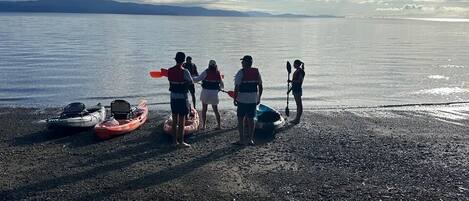A group of people on a pebbly beach with kayaks and a paddleboard, overlooking a calm body of water.