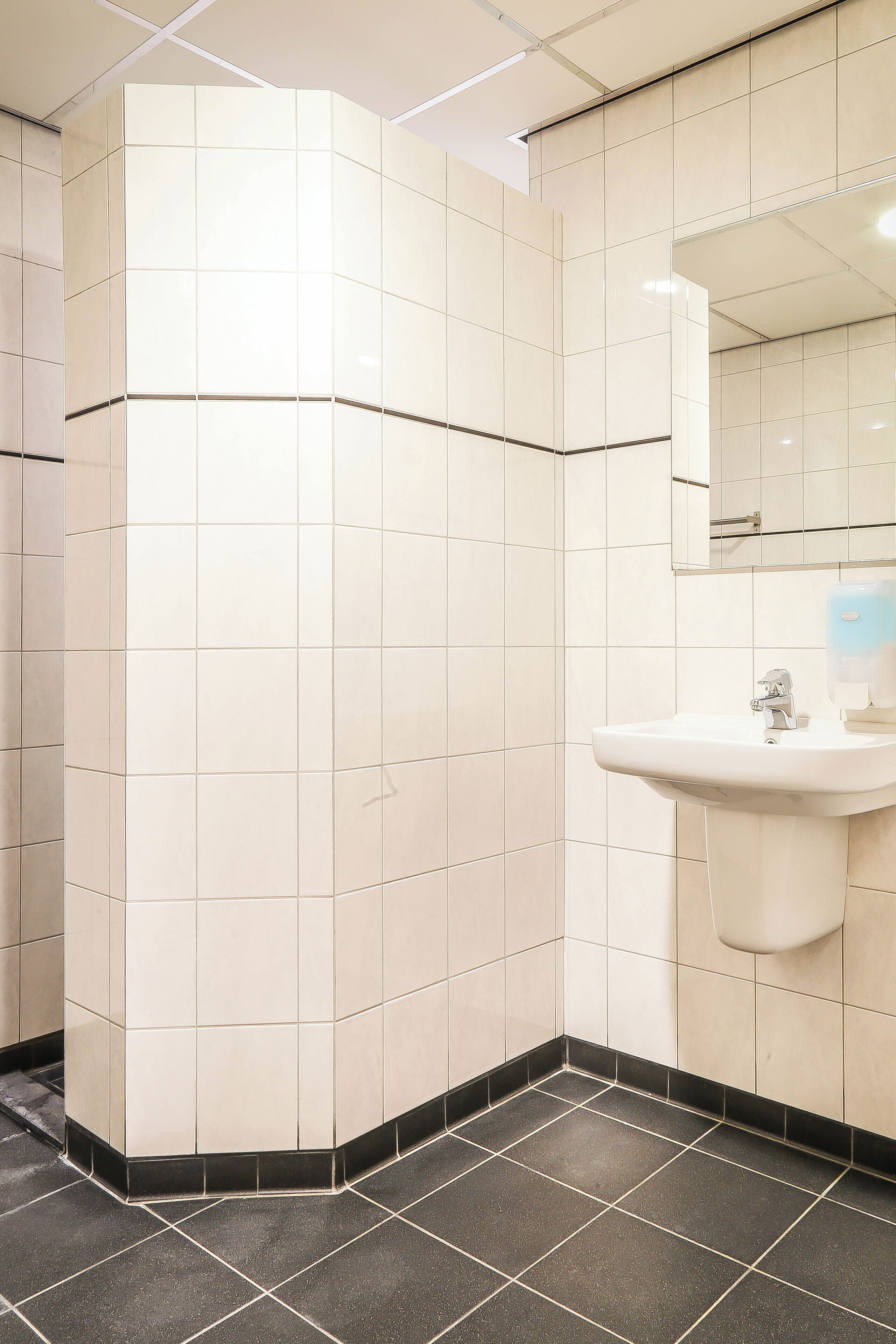 Un baño con paredes de azulejos blancos, piso de losetas negras, un lavamanos y un espejo.